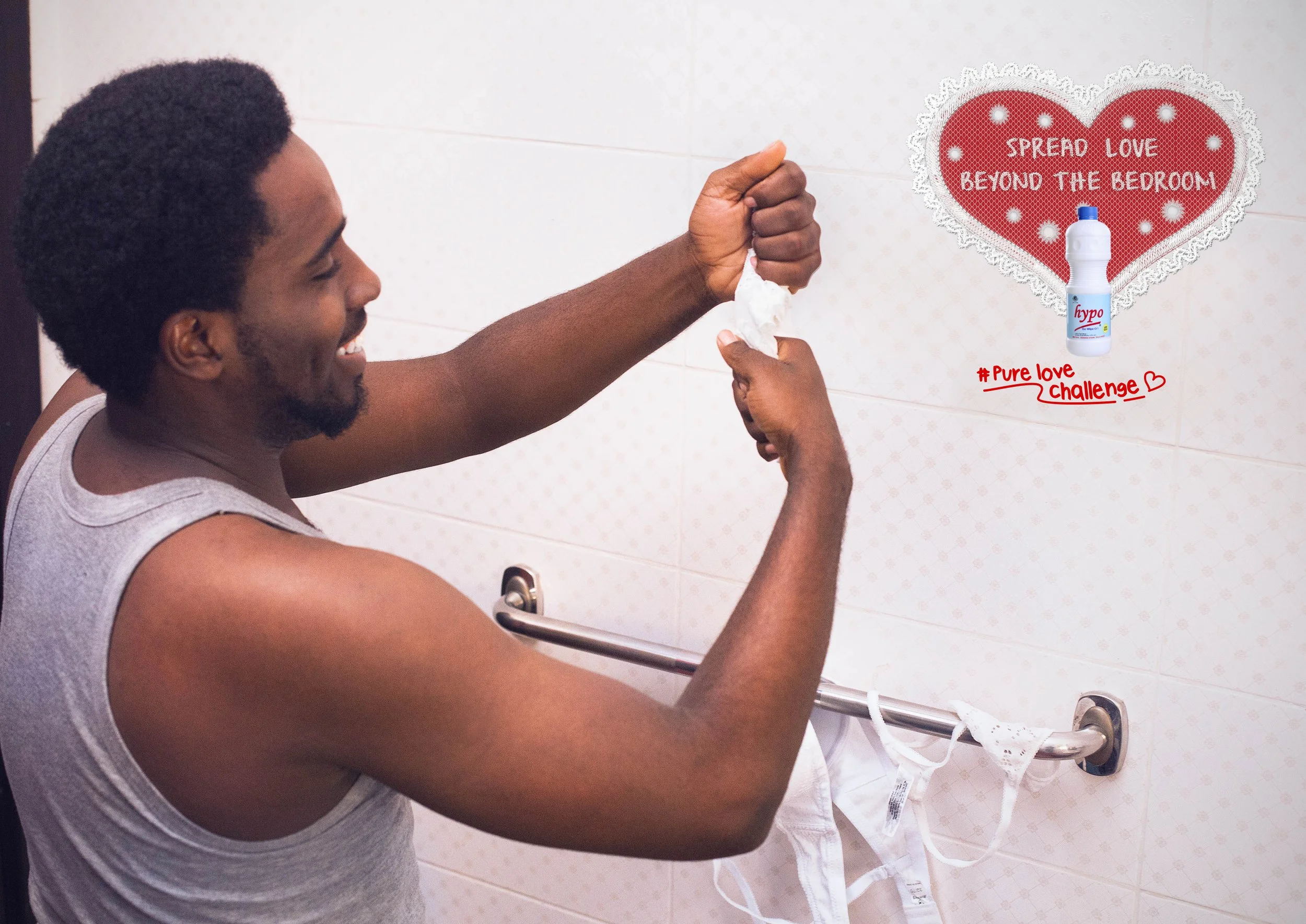 A man with short curly hair and a beard is smiling and holding a tissue in his right hand. He is standing in a bathroom, near a white tiled wall, with a grab bar and a face mask hanging from it. On the wall, there is a decorative heart-shaped sign that says, 'Spread love beyond the bedroom,' with a picture of a bottle of disinfectant spray and the hashtag '#PureLoveChallenge' written below.
