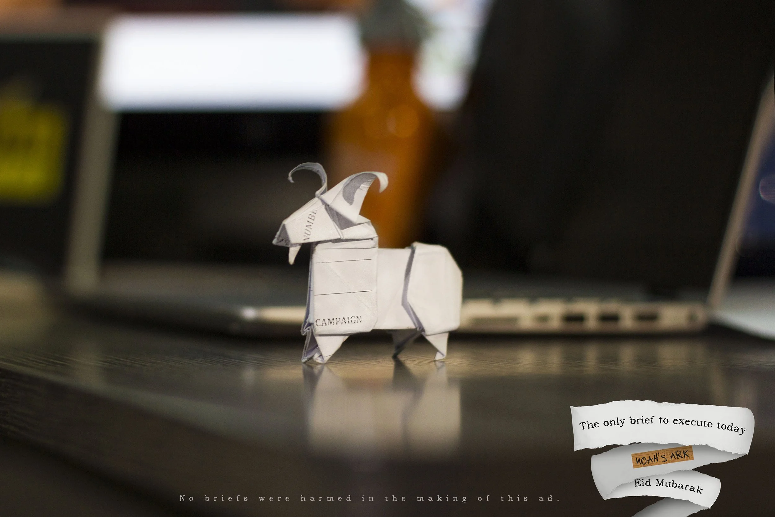 An origami elephant with a newspaper word on its body, placed on a desk with blurred monitors in the background. There are torn paper notes with phrases like 'The only brief to execute today' and 'Eid Mubarak' visible around the elephant.