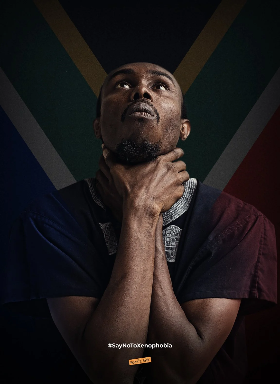 A man with short hair and a goatee looks upwards, clutching his neck with both hands, in front of a background featuring the South African flag, with the hashtag #SayNoToXenophobia at the bottom and a small orange sticker reading 'MOAH'S ARK'.