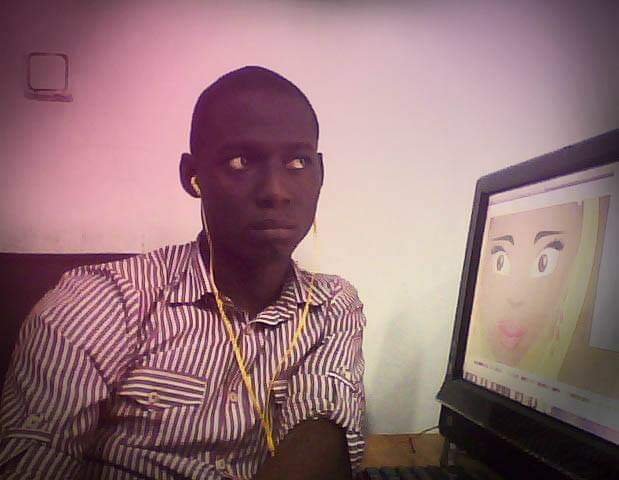 A young Nigerian art director with dark skin wearing a striped shirt, earbuds, and a yellow lanyard, sitting at a desk and looking at a computer monitor showing a cartoon face.