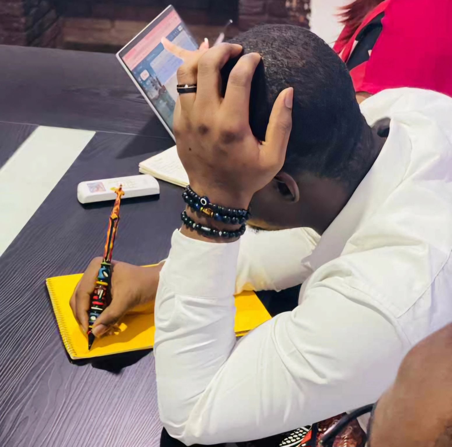 A person with short black hair wearing a white shirt is resting their head on their left hand while sitting at a table. They are writing in a yellow notebook with a colorful pen. The person's right hand is on the table, holding the pen, and they are wearing several black bead bracelets. A TV remote, an open notebook, and a small white object are on the table. A person wearing a red and black shirt is partially visible in the background.