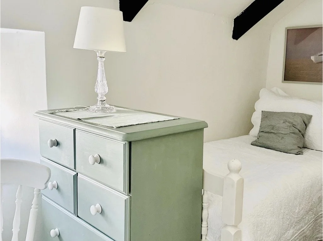 A bedroom corner with a white bed, a gray pillow, a wooden picture frame on the wall, a green dresser with four drawers, a white table runner, and a white lamp on top.