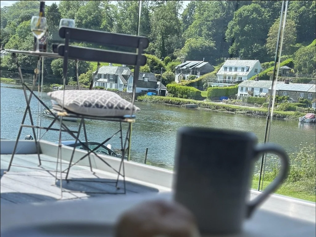 View of a river with several houses and lush greenery across the water; inside a boat, a metal rack with a pillow, a blurred mug, and a glass of drink are visible.