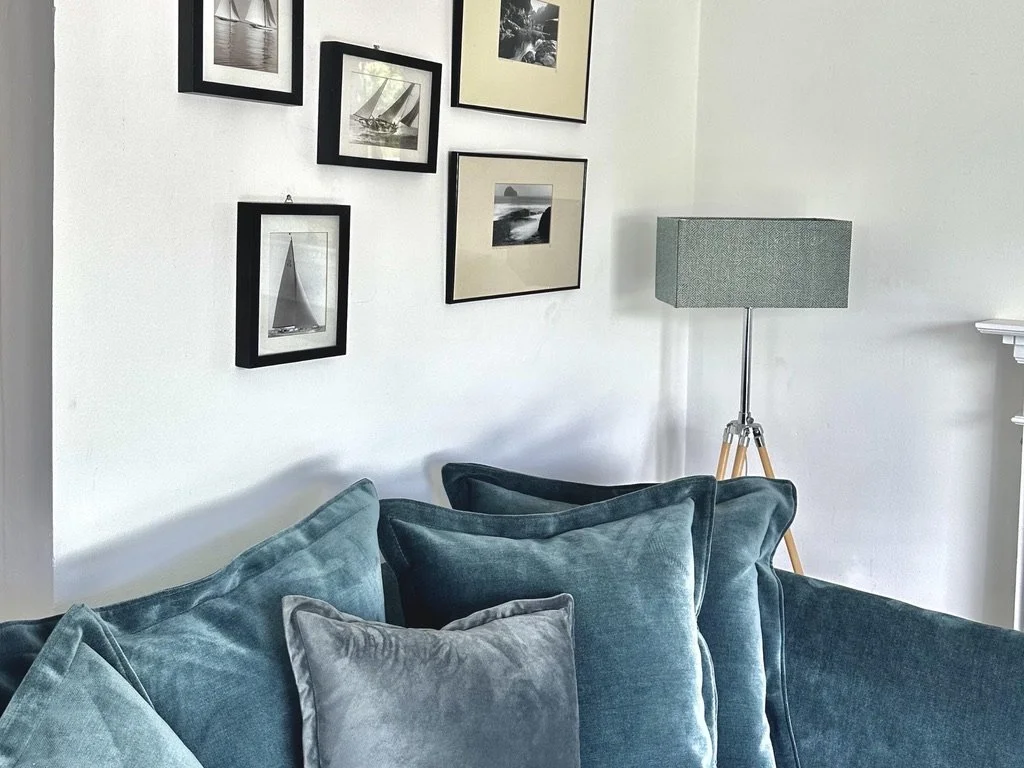 Living room corner with framed black and white photographs of sailboats and coastal scenes on the wall, a tall floor lamp with a gray shade and wooden tripod base, and a teal velvet couch with multiple cushions.