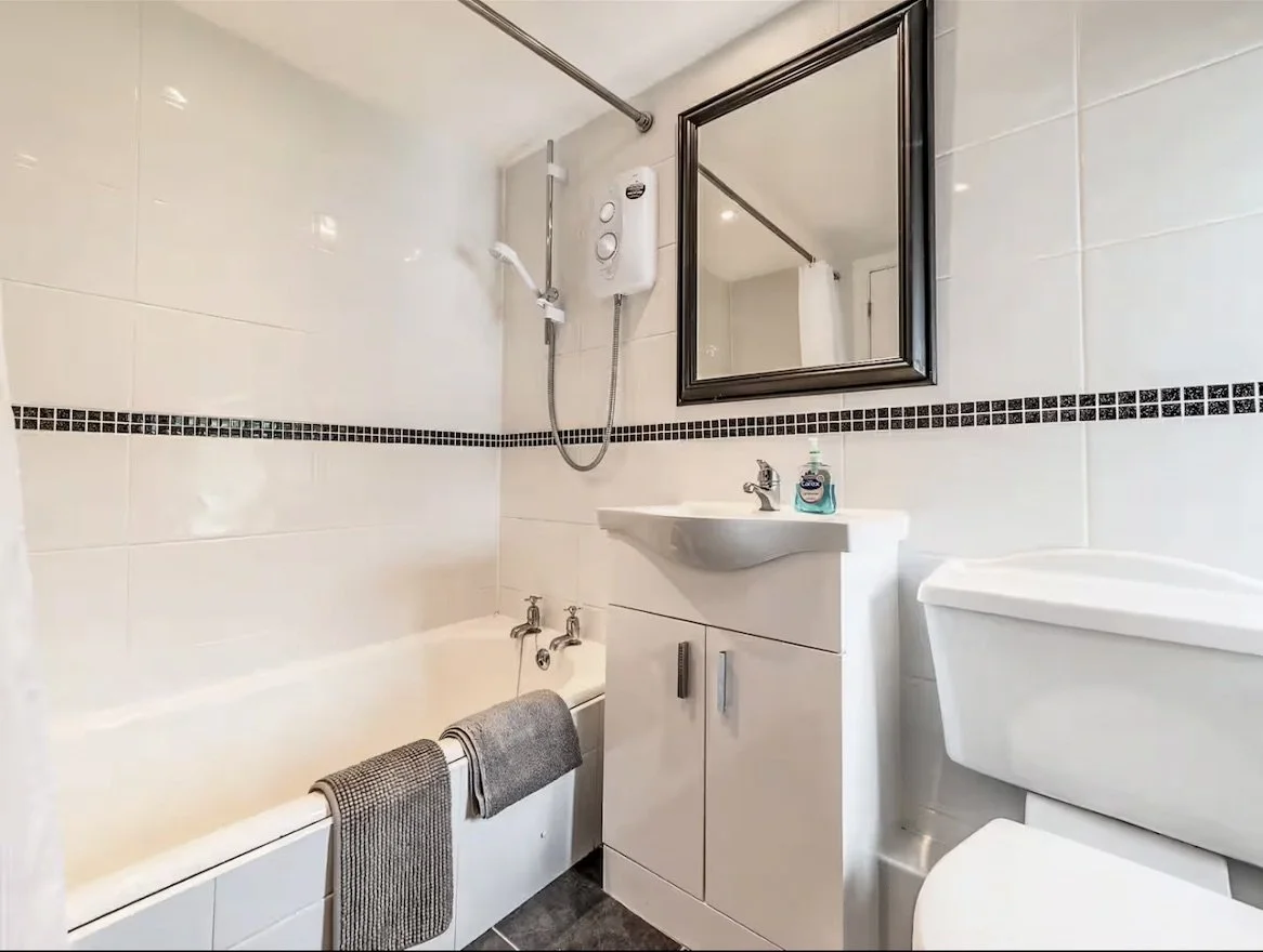 Small bathroom with a bathtub, sink with a cabinet, and a toilet. The bathtub has a hand towel draped over its side. The sink has a soap dispenser. The wall has a horizontal black tile border and a mirror above the sink. A shower head and water heate