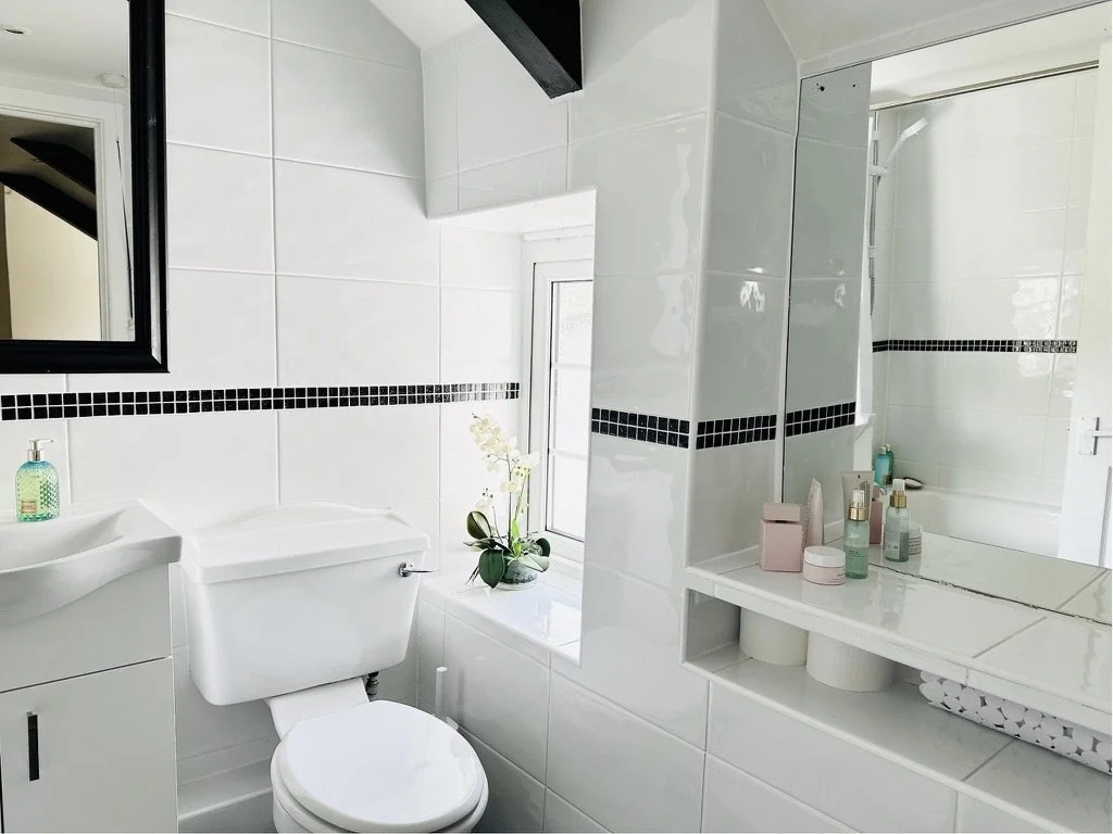A clean, modern bathroom with white tiles, a toilet, a window with a potted plant, a small sink with a green soap dispenser, and a mirror above the sink. There are various toiletries and containers on the windowsill and near the mirror.