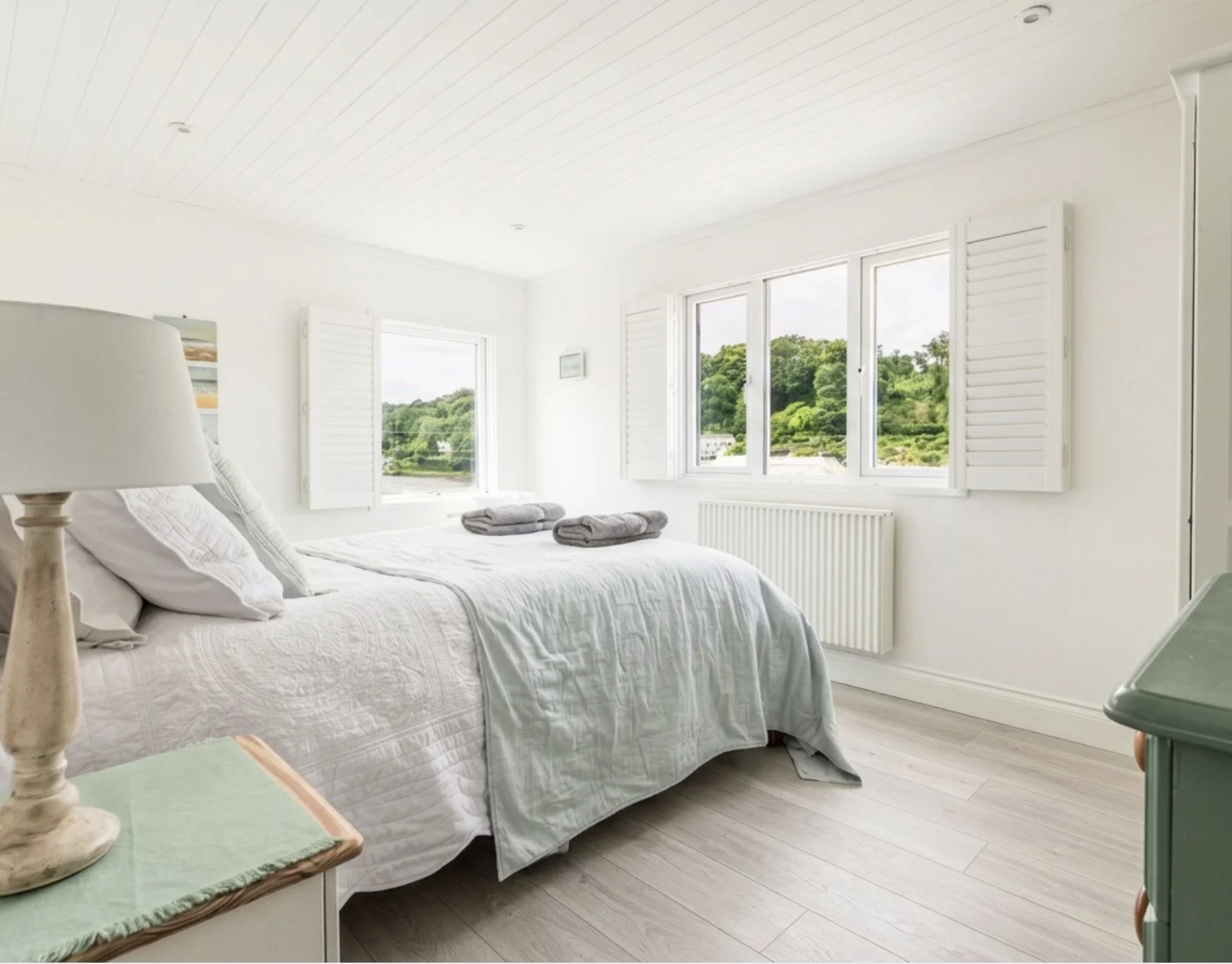 A bright bedroom with large windows showing green trees outside. The bed has white bedding, two pillows, and neatly folded towels on top. There is a bedside table with a lamp and a green cloth, and a green chest of drawers. The room has white walls a