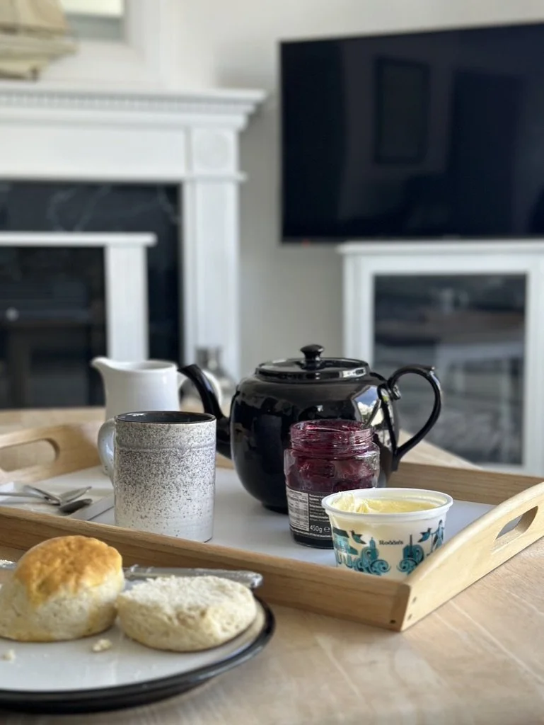 A breakfast tray with a teapot, cups, jam, and butter, placed on a wooden table in a cozy living room.