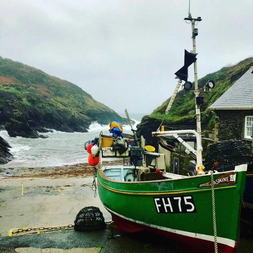 Green fishing boat with registration FH75 docked on a rocky shoreline near a building, with a hillside and rough sea in the background, overcast sky.