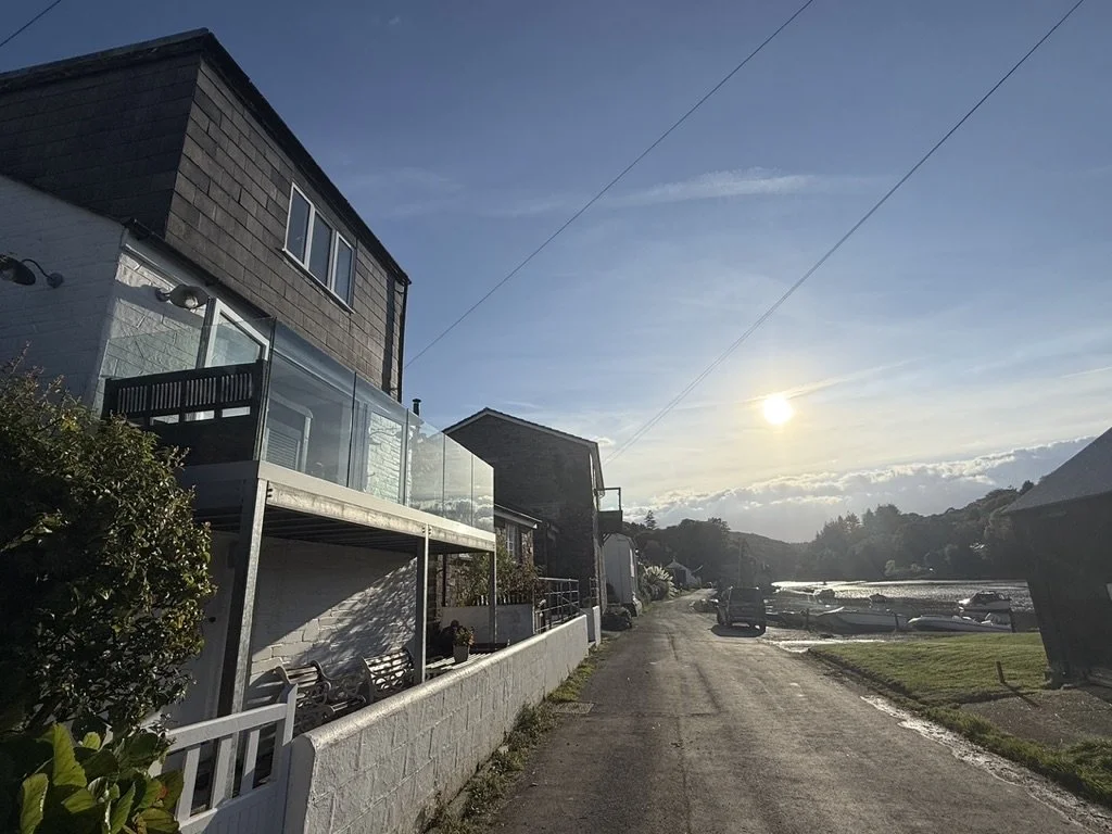 Residential street with houses, boats on water, and mountains in the distance under a setting sun.