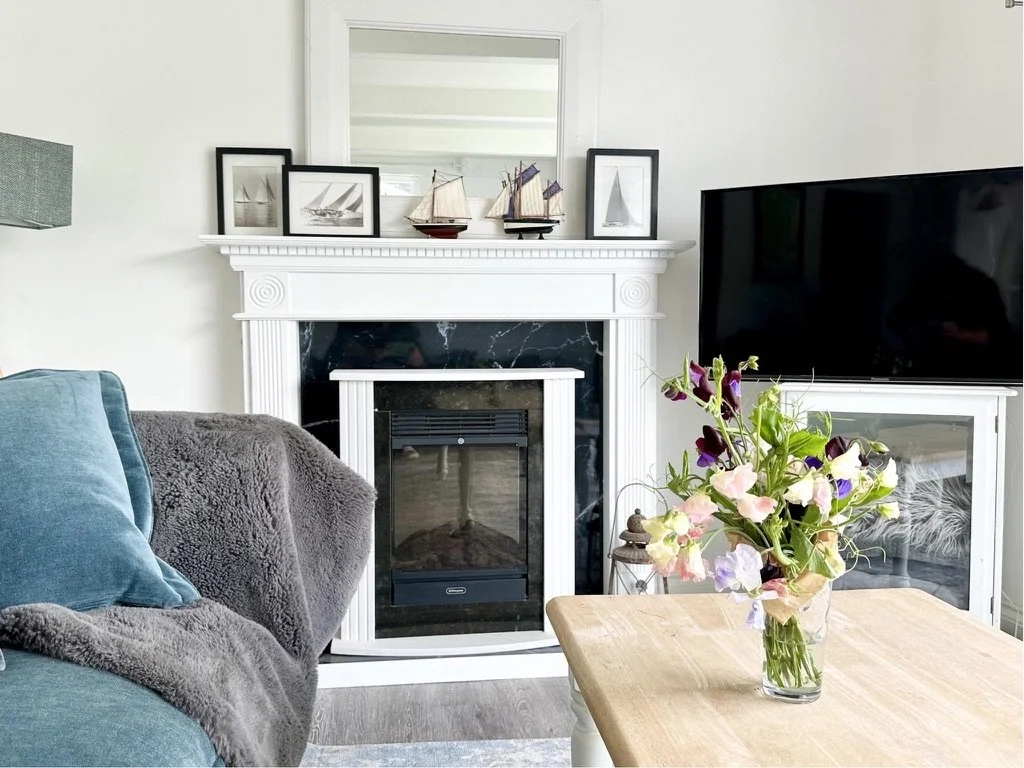 Living room with a fireplace, framed pictures, small ship models on fireplace mantel, a TV, a side table with a lantern, and a table with a vase of flowers.