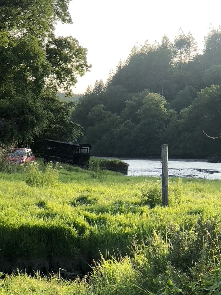 Green grassy area next to a river with trees and hills in the background, and a vehicle and a black structure near the water.
