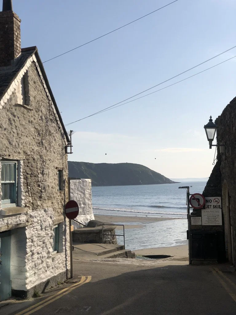 View through a narrow street leading to a beach with calm water and a hillside in the background. On either side of the street are stone buildings, with signs indicating no entry and no jet skis.
