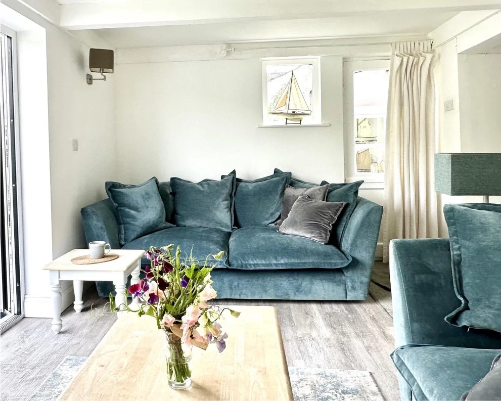 Living room with teal velvet sofa, a white side table, a coffee table with a flower vase, and a sailboat decoration on the window ledge.