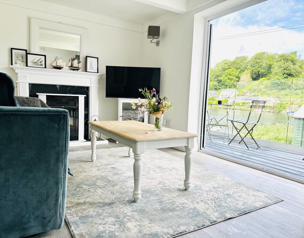 Living room with a fireplace, a flat-screen TV, a wooden coffee table with a vase of flowers, and a sliding glass door leading to a balcony with outdoor seating and a scenic view of green trees and water.