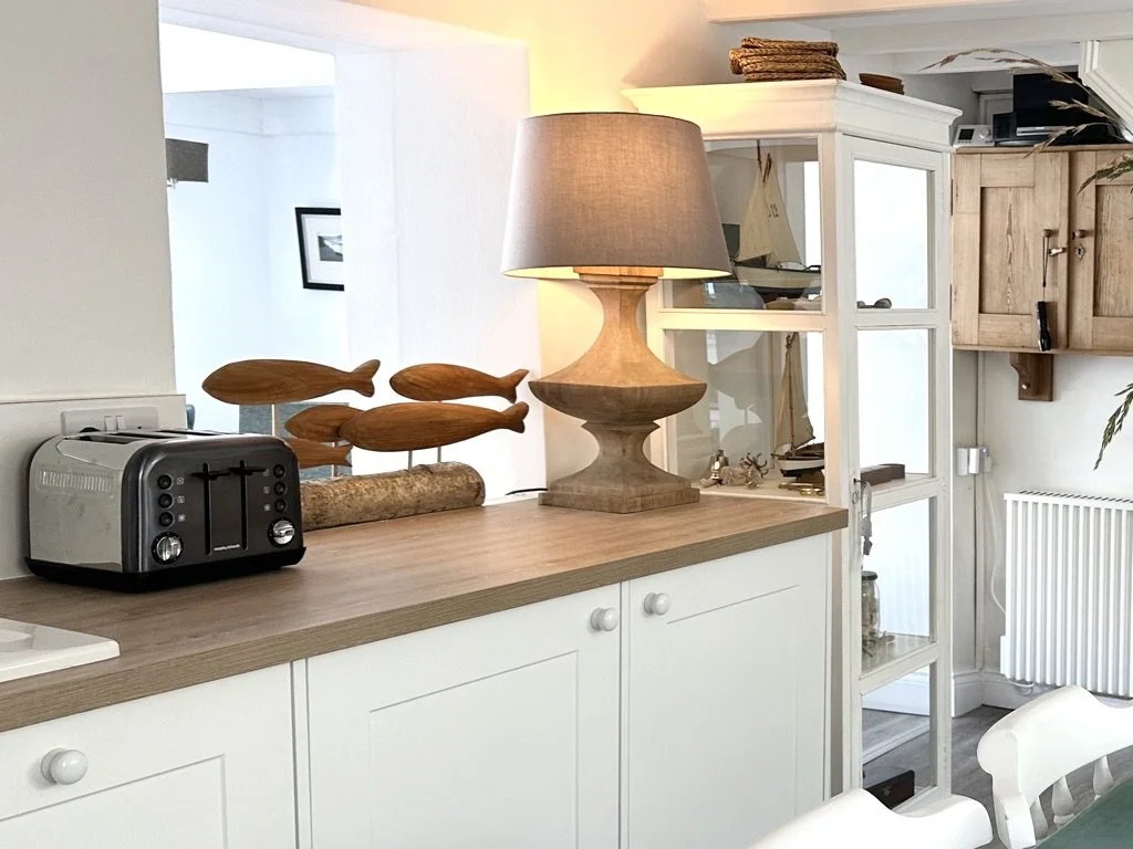A wooden countertop with a toaster, a wooden fish sculpture, and a table lamp with a cloth shade. Behind is a white open shelving unit with nautical decorations and a wooden cabinet, in a cozy kitchen or dining area.