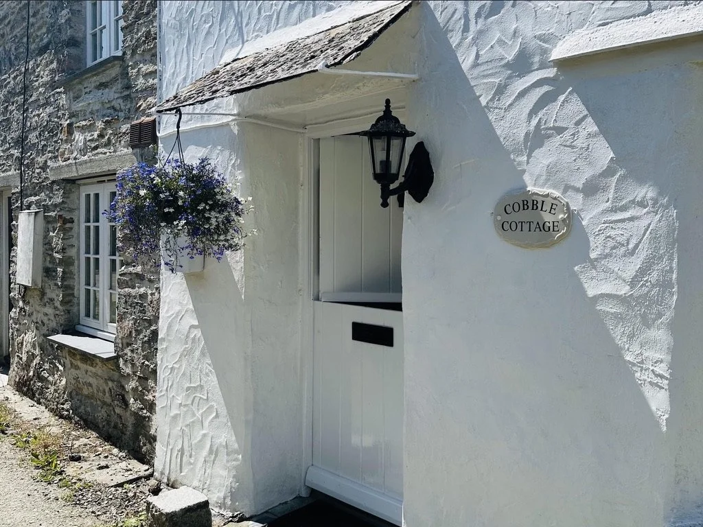 White cottage with hanging flower basket, black lantern light, and a sign that reads "Cobble Cottage."
