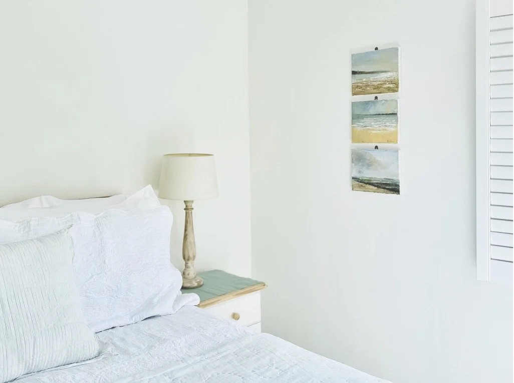 A bedroom corner with a bed, white bedding, a nightstand, a lamp, and three landscape paintings on the wall.