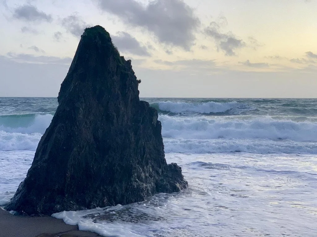 A large dark rock formation on the beach with ocean waves crashing against it, under a cloudy sky during sunset or sunrise.