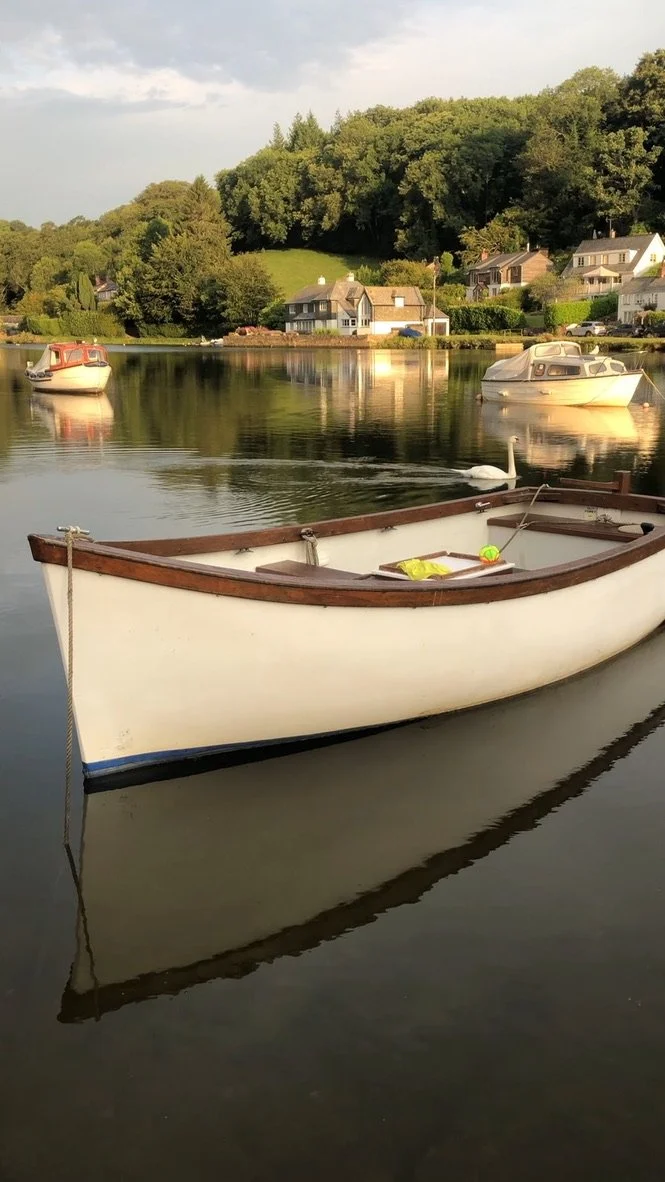 A white rowboat with a wooden trim floating on calm water, with two other boats and houses on a hill in the background.