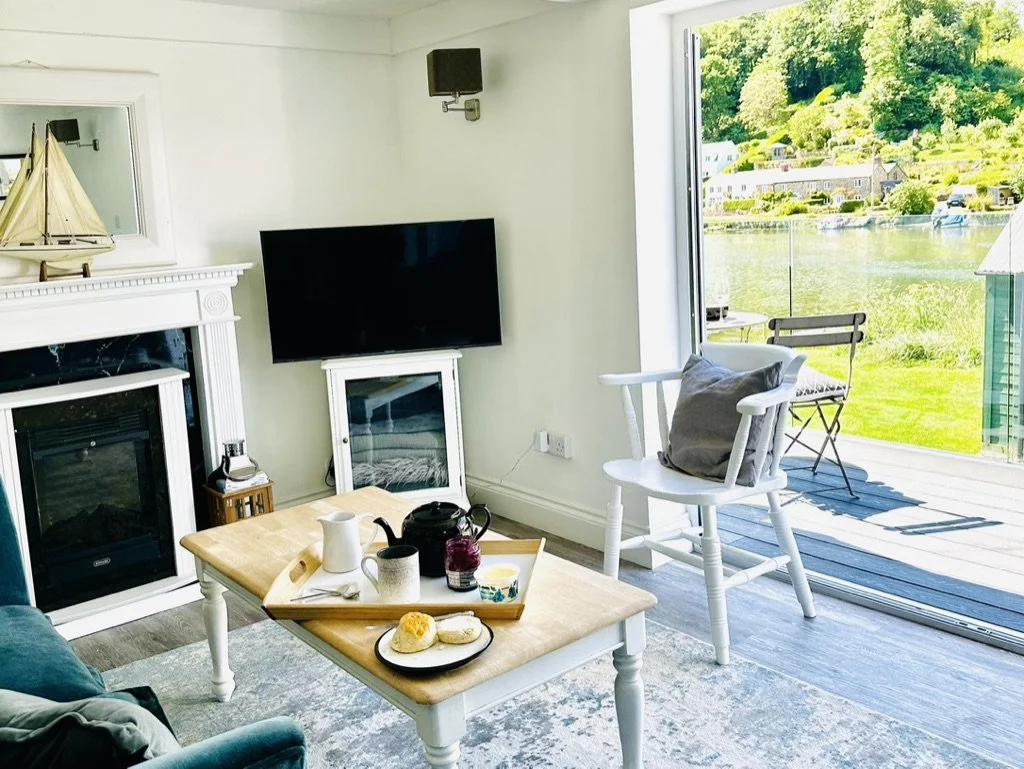 Living room with a fireplace, a flat-screen TV, a coffee table with snacks and drinks, a white rocking chair with a pillow, and a sliding door opening to a deck overlooking a lake with houses and greenery.