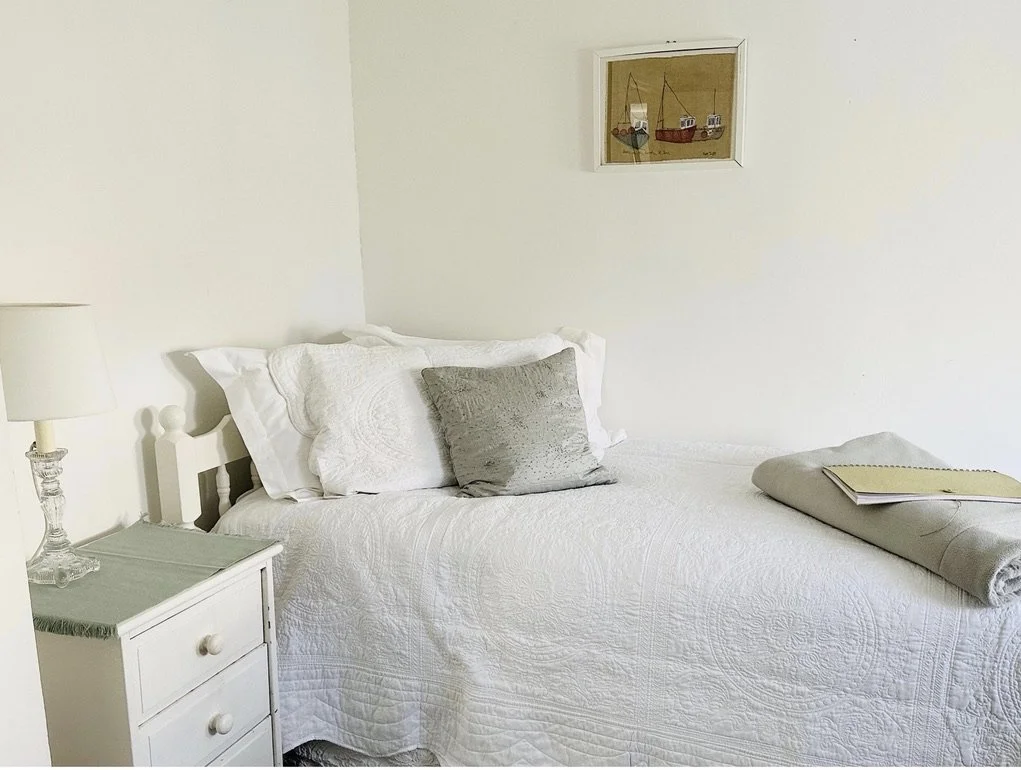 A neatly made bed with white bedding, two pillows, and a gray cushion in a bedroom. A white nightstand with a glass lamp and books, and a small framed picture of boats on the wall.