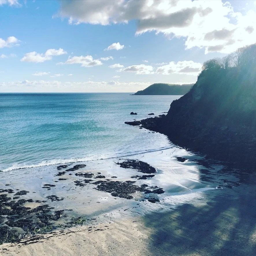 View of a rocky sandy beach with turquoise ocean water and a hill in the distance under a partly cloudy sky.