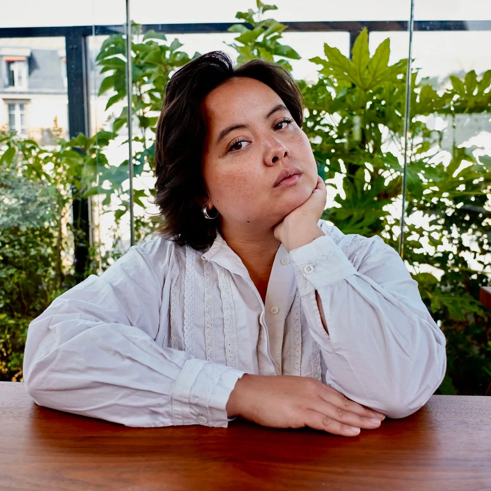 L'image montre Louise Feld, une artiste contemporaine émergente qui réalise des tapisseries et du contenu pour les réseaux sociaux. Elle s'appuie sur un comptoir, et pose sa tête sur sa main. Végétation derrière. Photo prise au Terrass Hotel, Paris