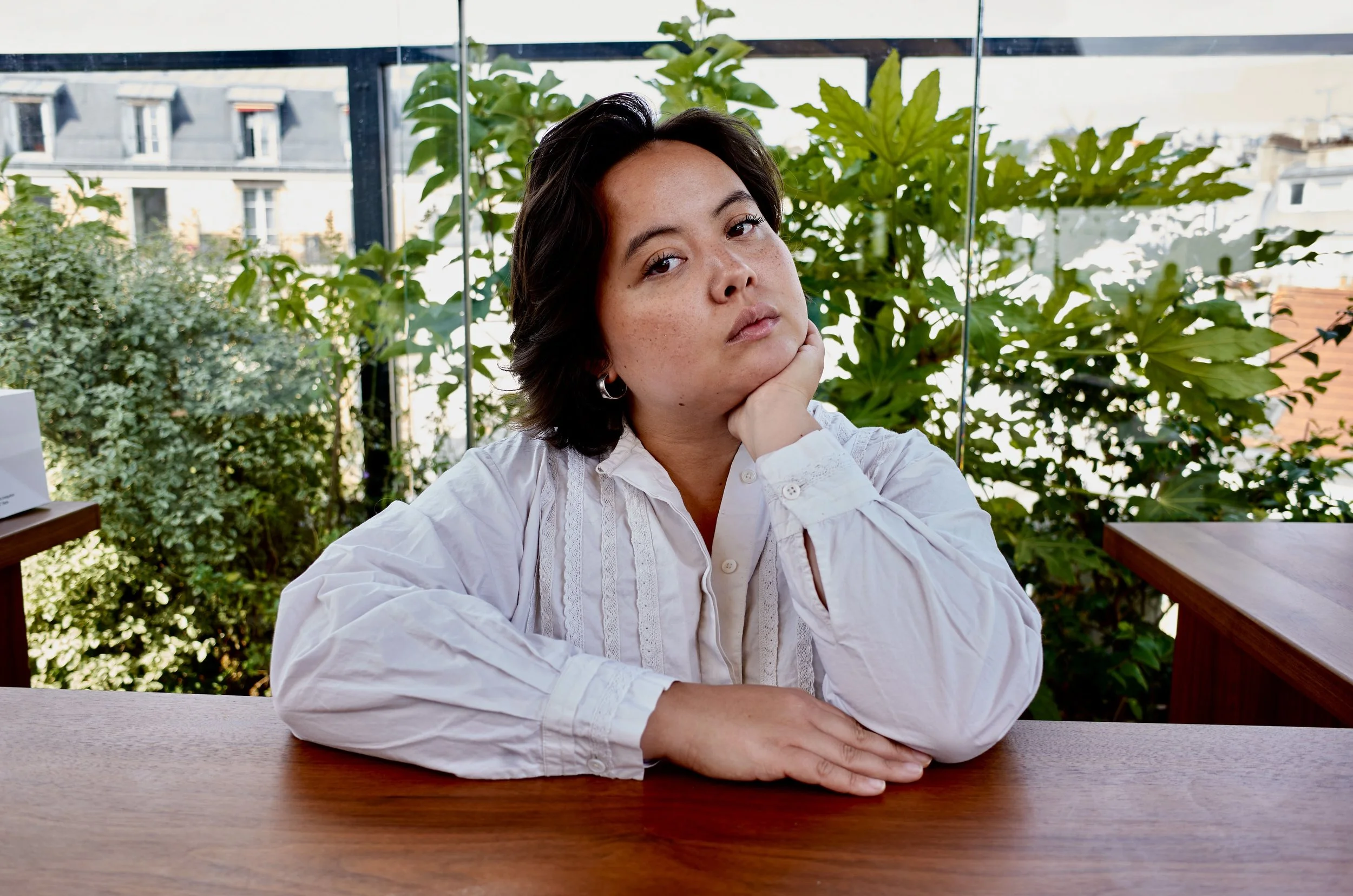 L'image montre Louise Feld, une artiste contemporaine émergente qui réalise des tapisseries et du contenu pour les réseaux sociaux. Elle s'appuie sur un comptoir, et pose sa tête sur sa main. Végétation derrière. Photo prise au Terrass Hotel, Paris