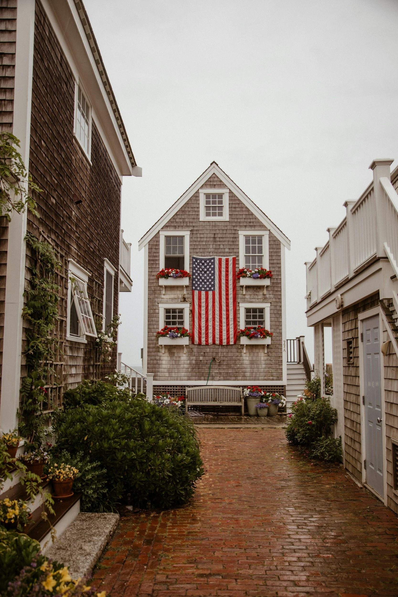 A narrow brick pathway leading to a house with a large American flag hanging from the second story, decorated with red, white, and blue flowers on the balconies, flanked by wooden buildings and greenery.