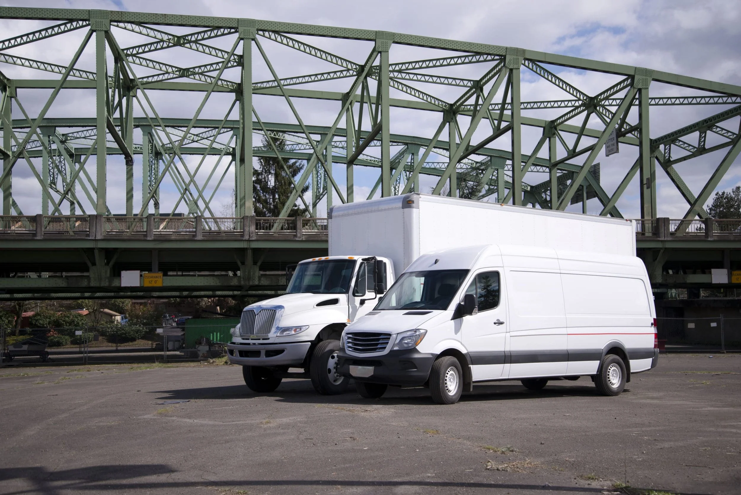 Two white delivery vans parked in a lot underneath a green metal bridge. Overcast sky with some clouds and visible trees in the background.