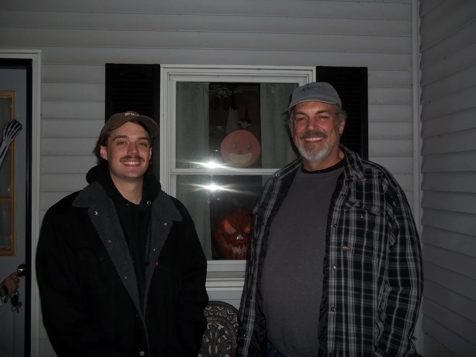 Two smiling men standing outside in front of a window with Halloween pumpkin decorations and a smiling face in the background.