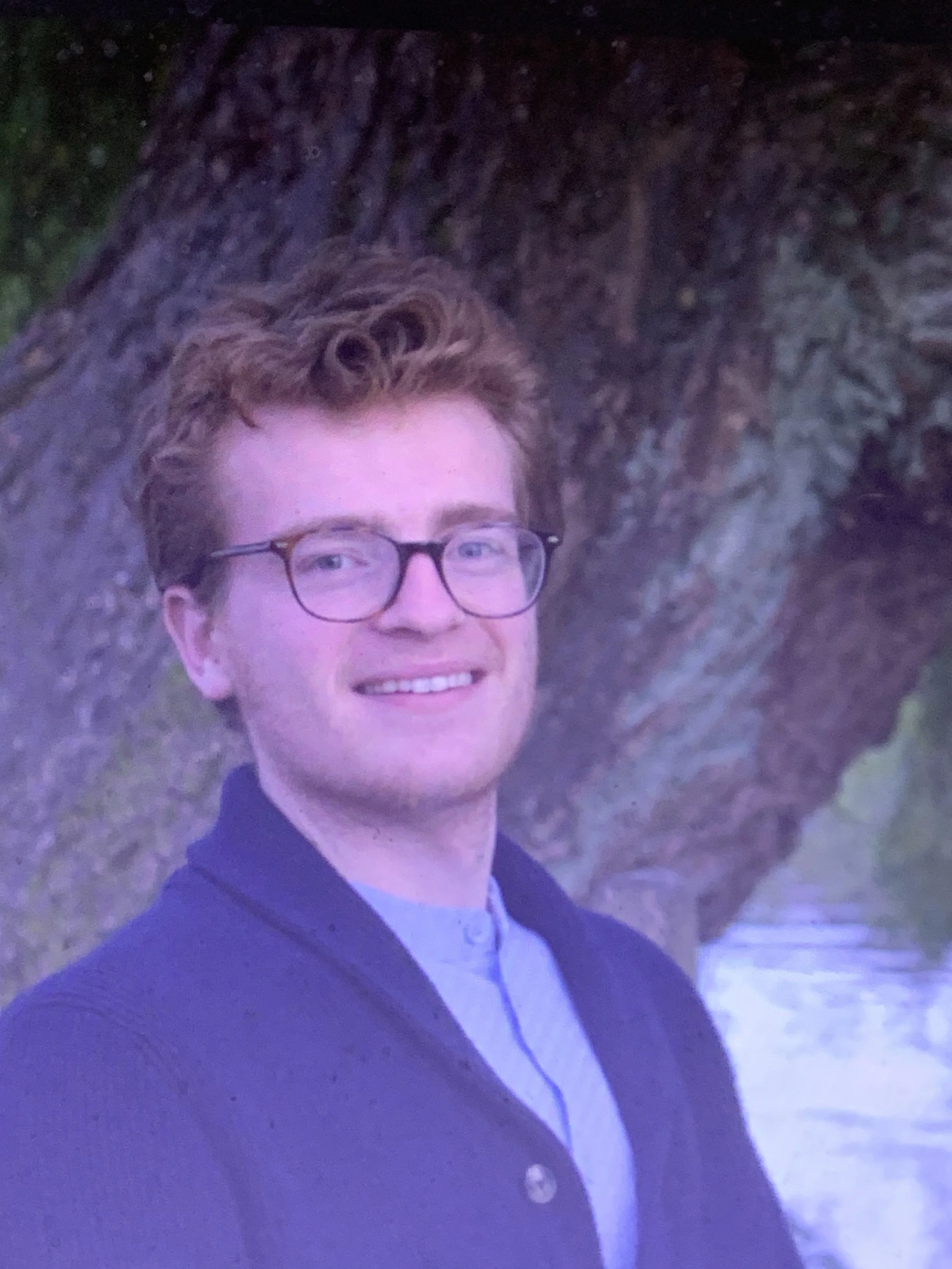 A young man with curly hair and glasses smiling outdoors in a dark setting.