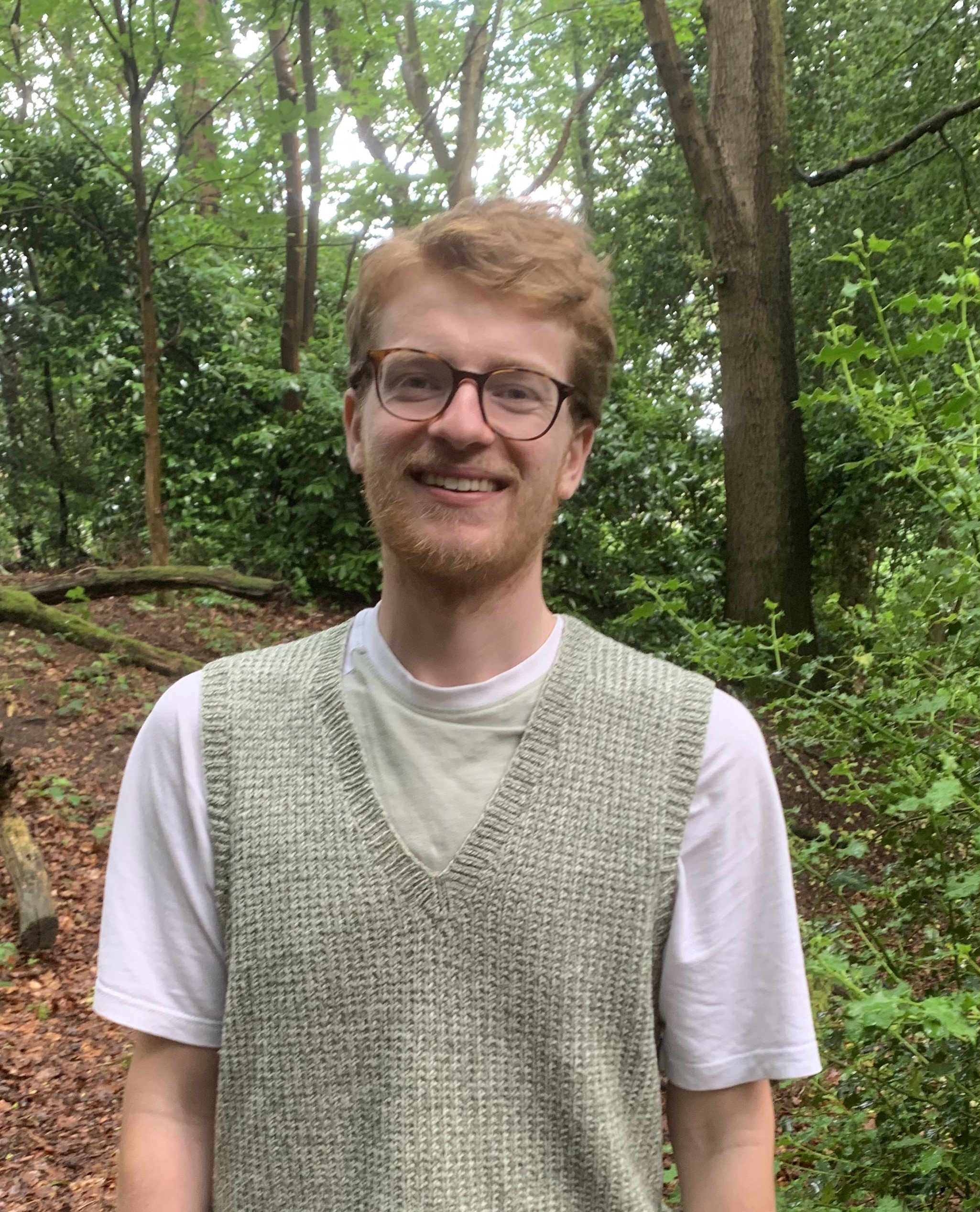 A young man with glasses and red hair smiling in a wooded outdoor setting.