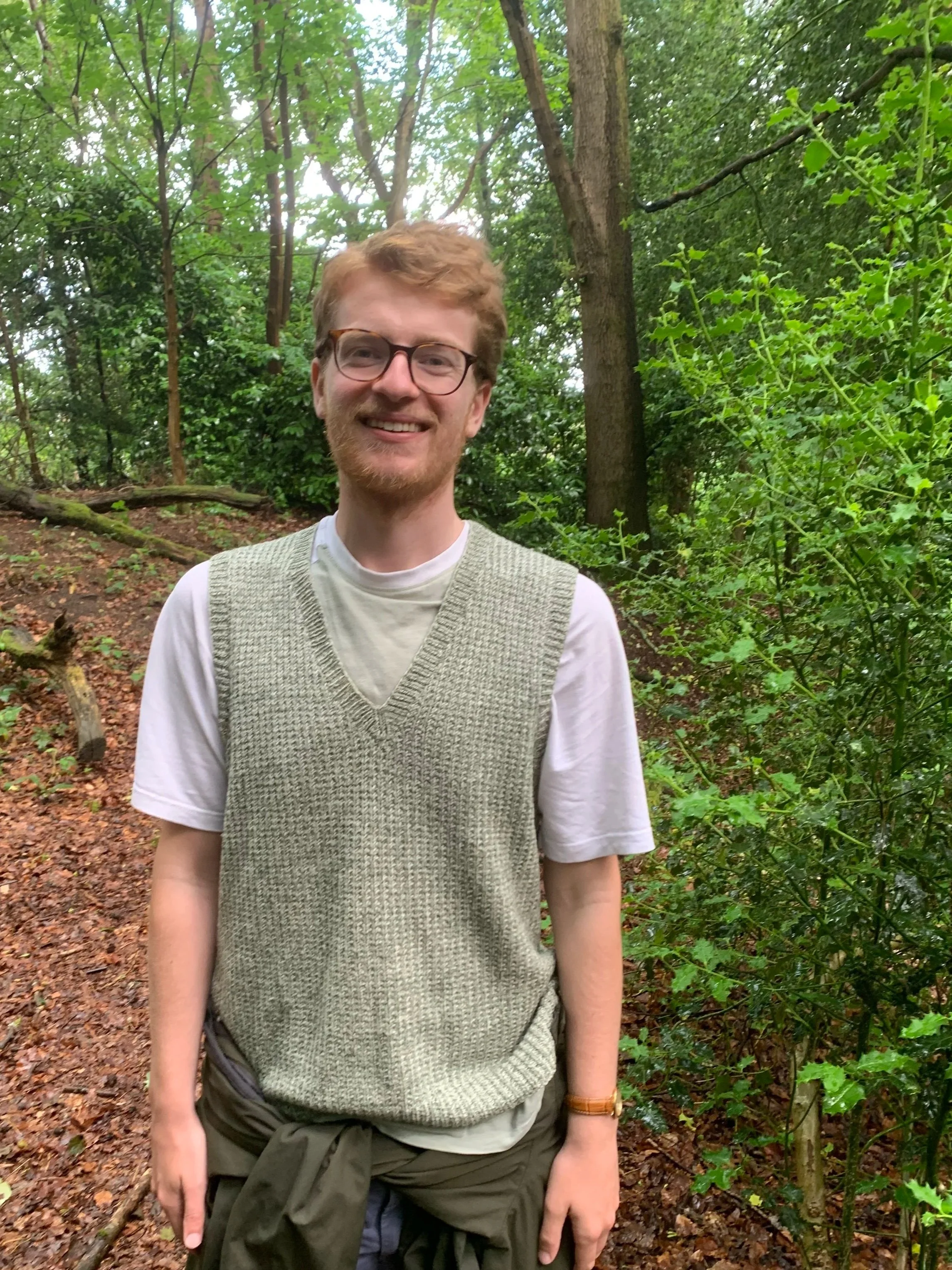A smiling young man with glasses and light brown hair in a forested area with dense green trees and shrubs, wearing a white t-shirt and a beige knitted vest, standing on a trail.