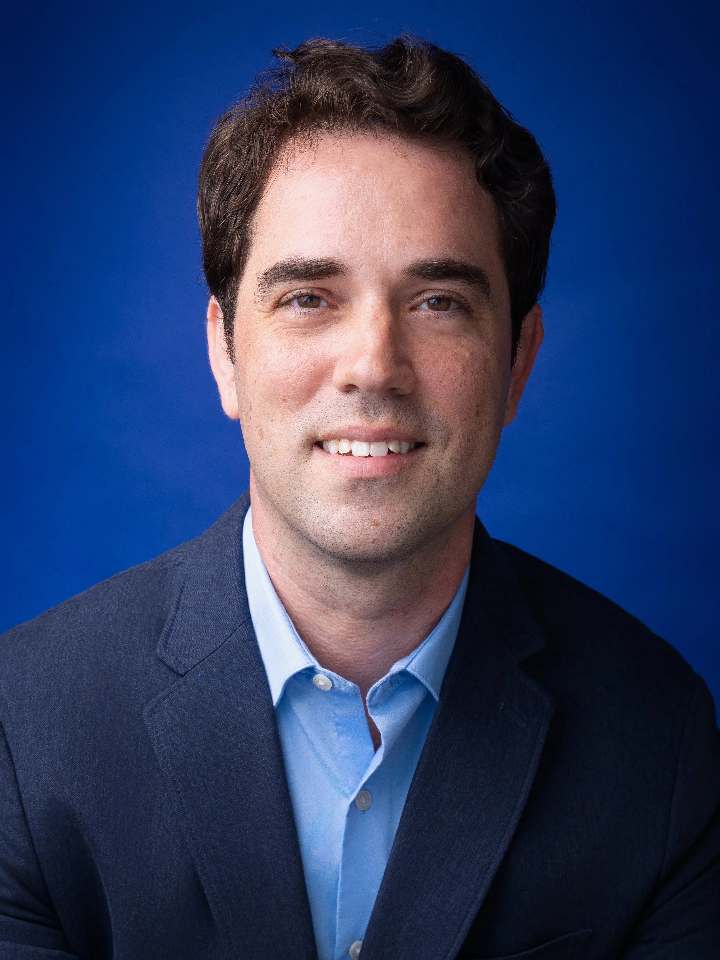Headshot of a man wearing a dark suit and light blue shirt, smiling, with a blue background.