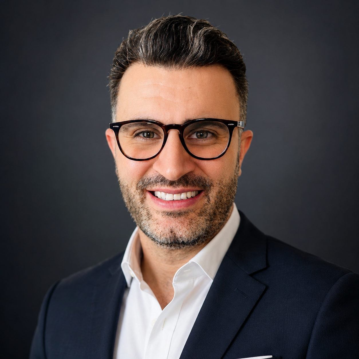 Headshot of a smiling man with dark hair, glasses, and a beard, wearing a dark suit and white shirt, against a dark background.