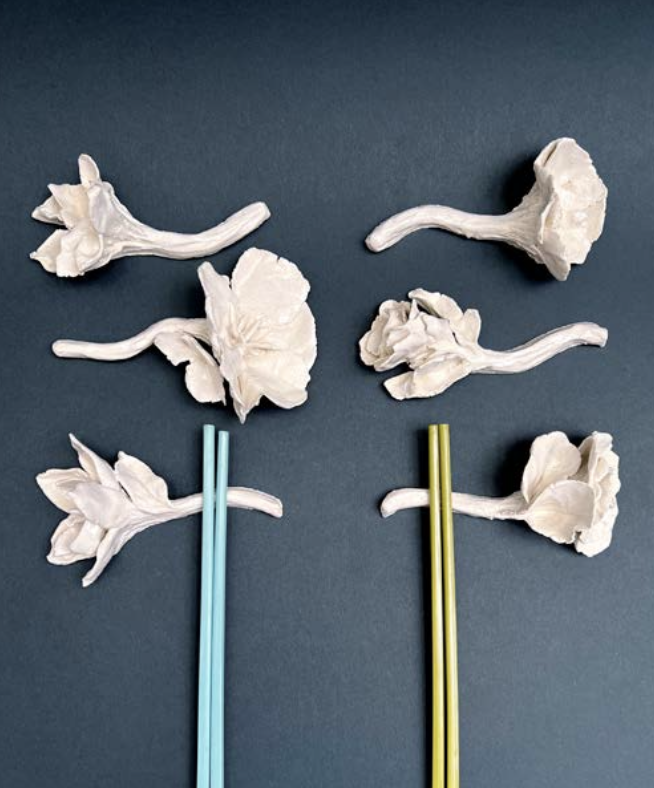 Six morceaux de fleurs en céramique ou plastique et deux pailles, une bleue et une jaune, disposés sur un fond gris.