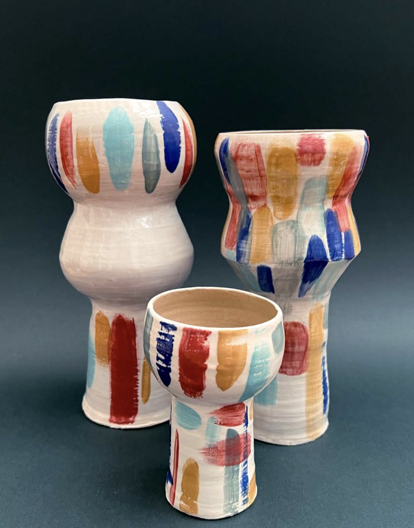 Vases en céramique colorés avec des motifs de taches rouges, bleues, jaunes et vertes sur un fond blanc, posés sur une surface sombre.