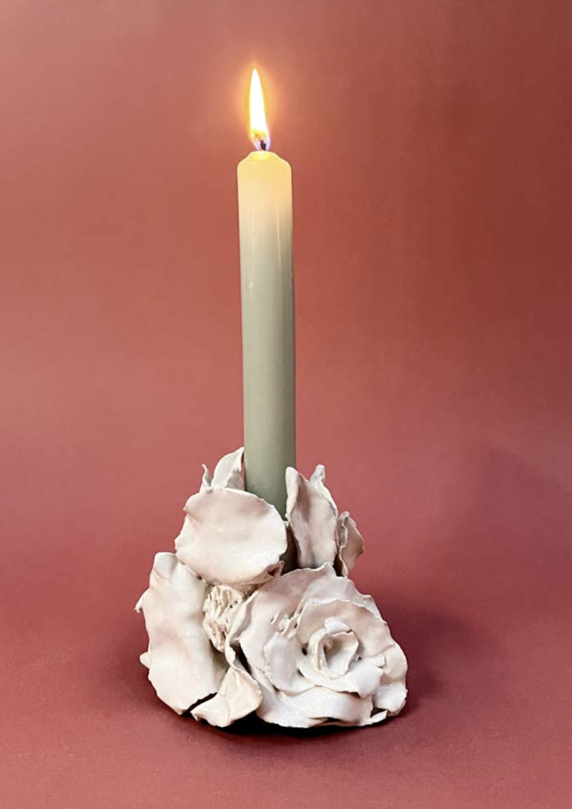 Une bougie blanche allumée posée sur un support décoratif en forme de roses en céramique blanche, avec un fond rose