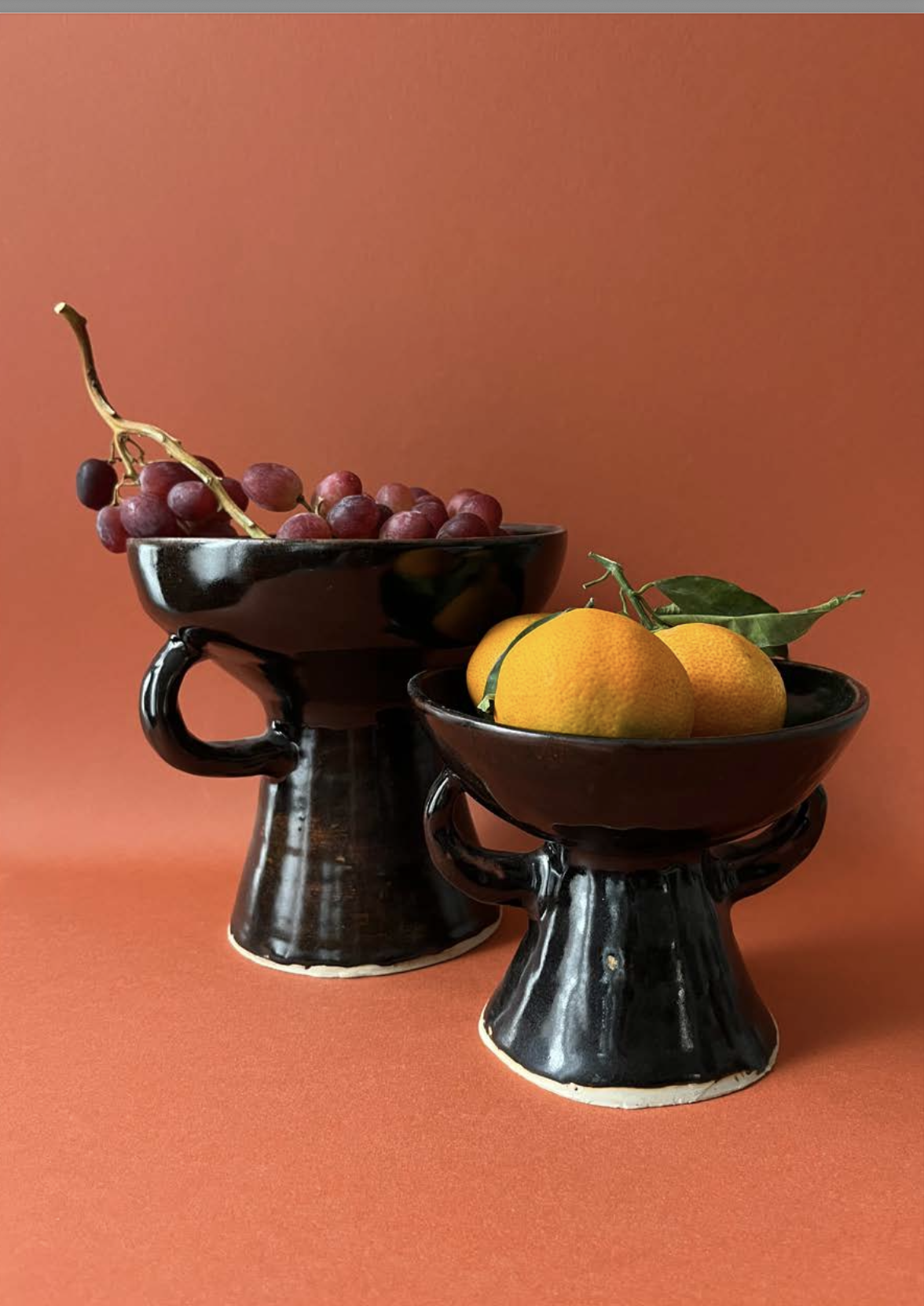 Deux coupes en céramique noire contenant des fruits, une avec des raisins rouges et une autre avec des oranges, sur un fond orange