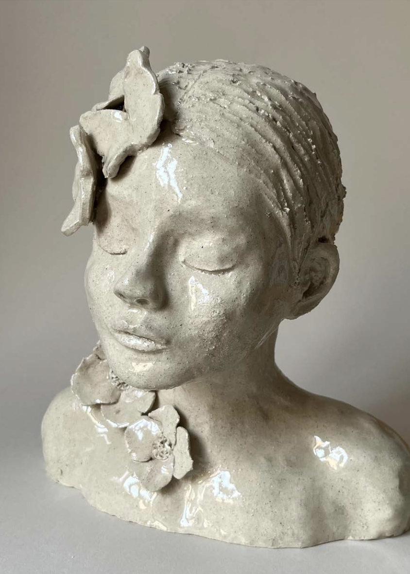 Buste en céramique d'une femme avec des détails floraux et un nœud dans ses cheveux, visage paisible avec les yeux fermés.