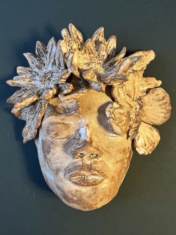 Masque en relief représentant un visage féminin avec des éléments floraux ou végétaux formant la coiffure, en tons dorés et argentés.
