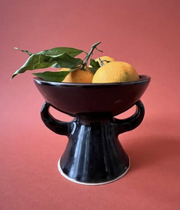 Un bol noir sur un support noir contenant des citrons et des feuilles vertes contre un fond rose.