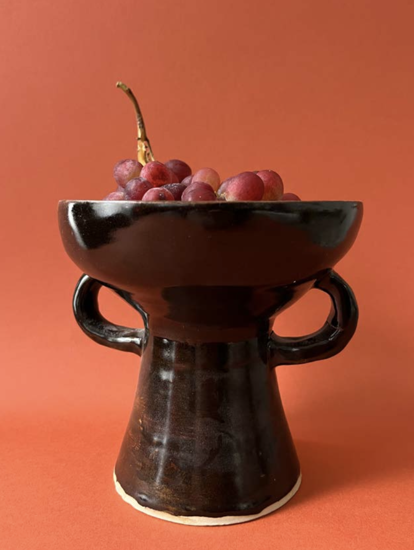 Une coupe noire contenant des grappes de raisin rouge, placé sur un support en céramique noire avec deux anses, contre un fond orange.