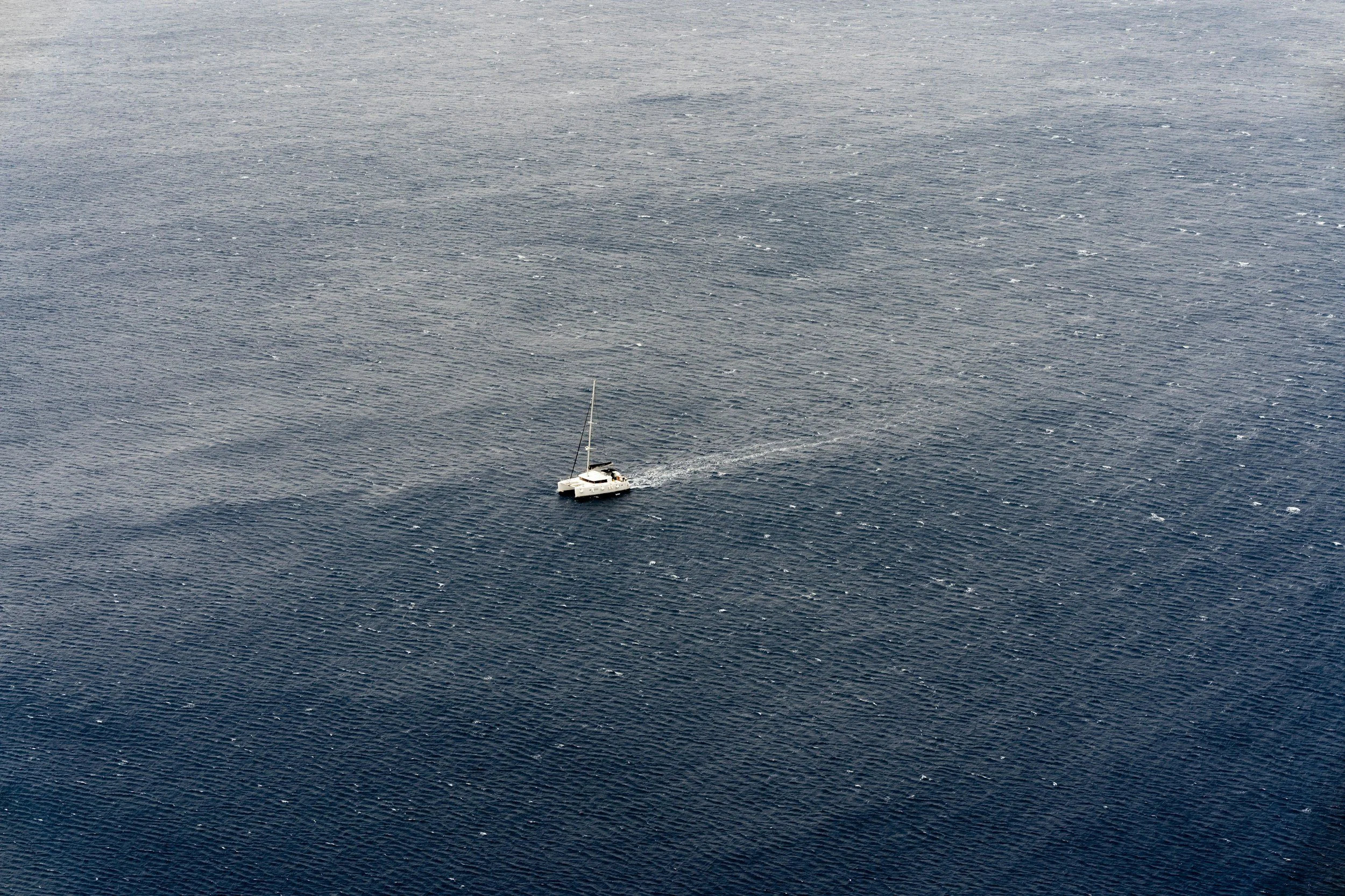 A small sailboat with a white hull and black sails sailing across a large body of water with rippling waves.