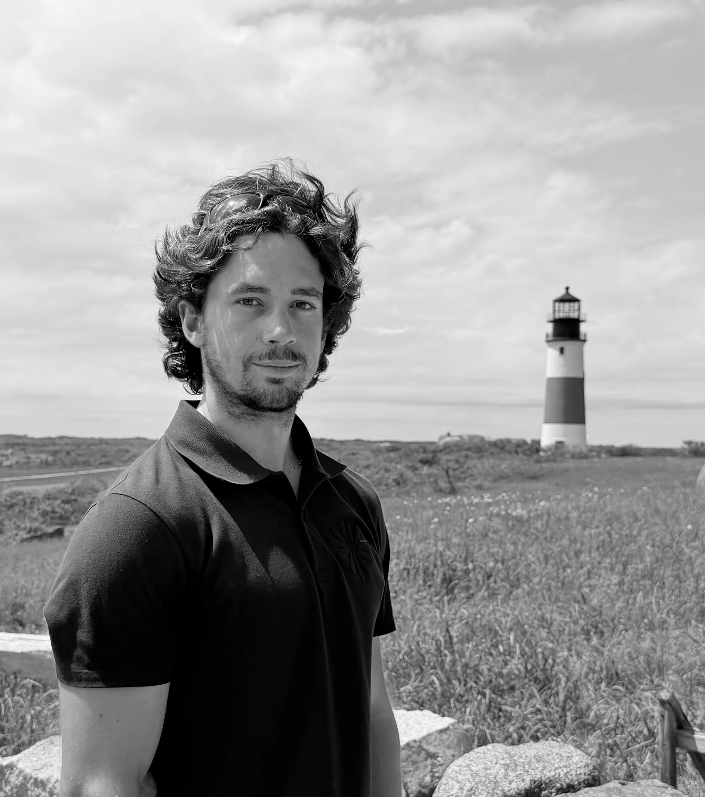 A man with curly hair and sunglasses resting on his head stands outdoors near a lighthouse, with a cloudy sky in the background.