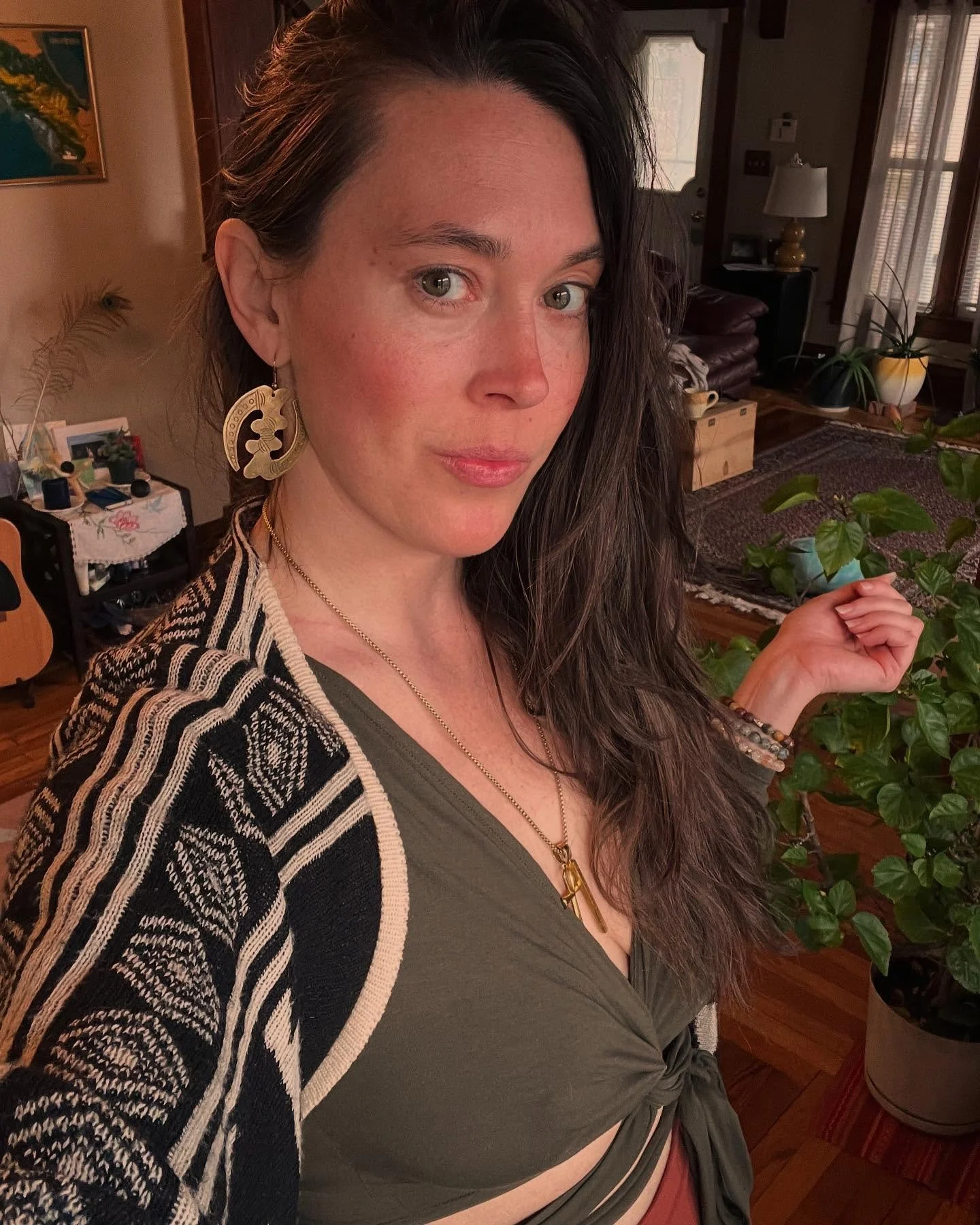 Meaghan Bergman, the president of The Healing Feast Foundation Board of Directors, is a woman with long dark hair and earrings looking at the camera, standing in a living room with plants, wooden furniture, and a patterned rug.