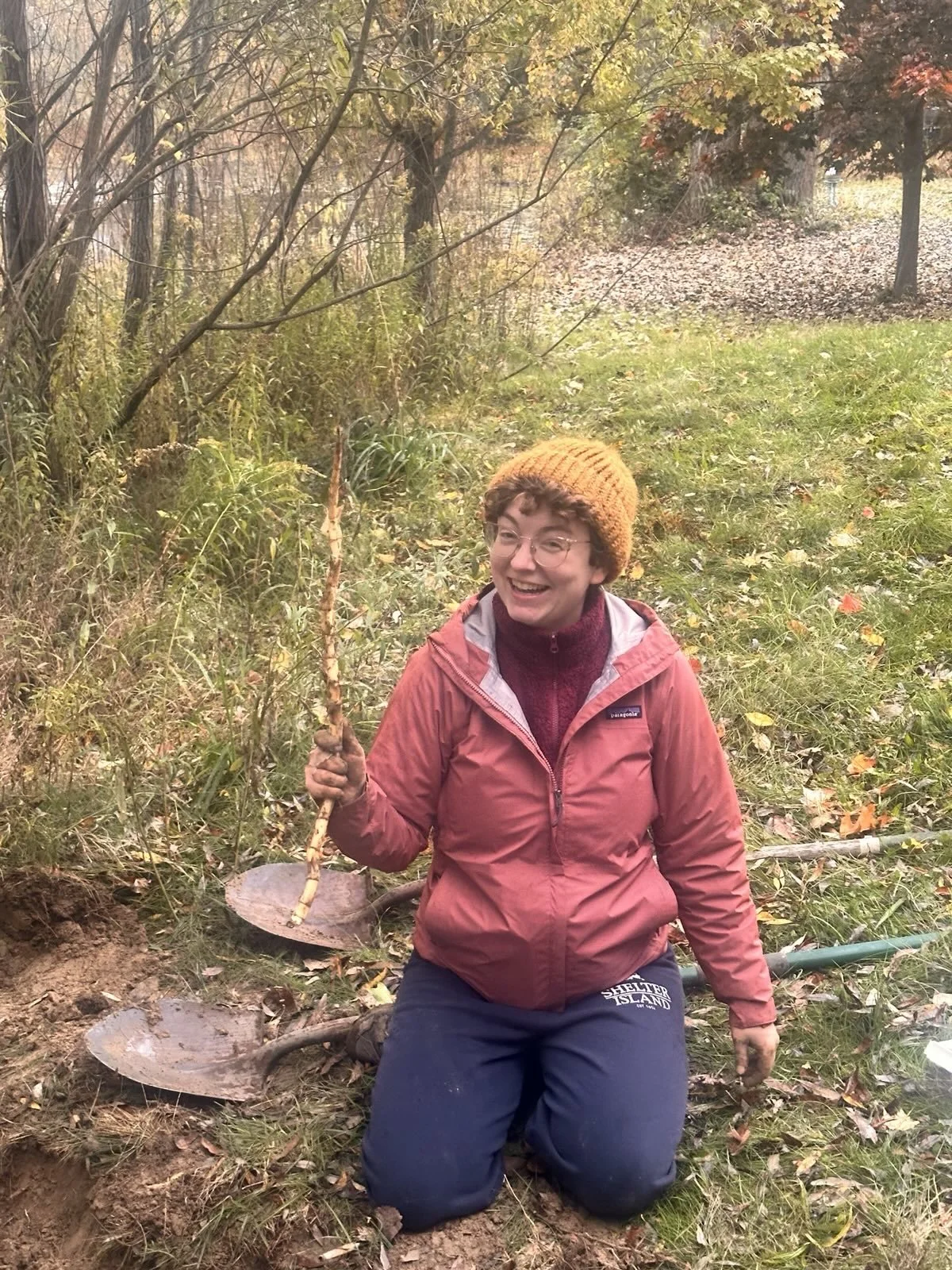 Dinah DeWald, member at large of The Healing Feast Foundation board, is a woman with glasses, a yellow knit hat, and a red jacket kneeling on the ground holding a plant with a bare root in one hand, outdoors surrounded by trees and fallen leaves.