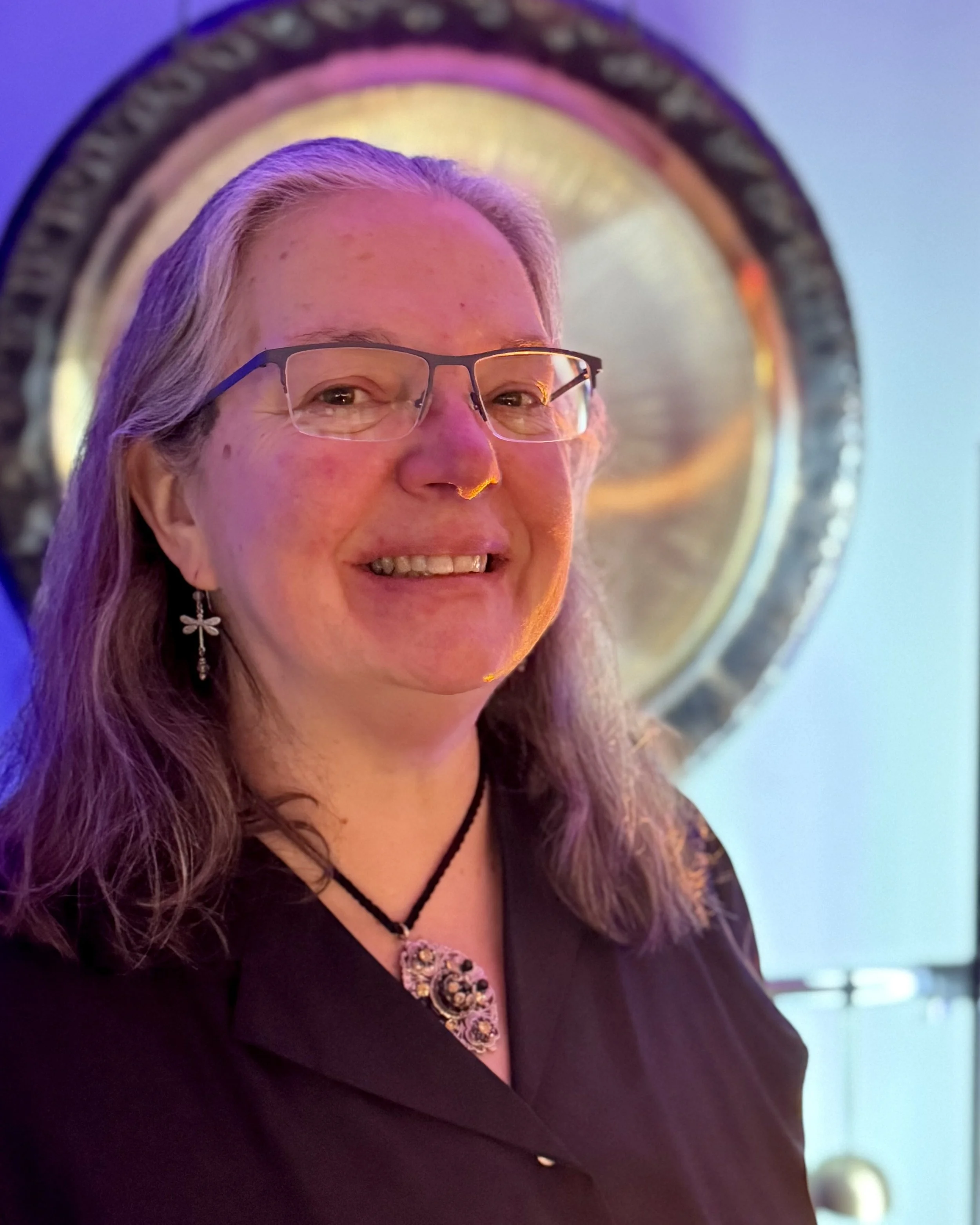Janice Leach, member at large of The Healing Feast Foundation, smiles at the camera standing in front of a a silver disc with blue-ish lighting
